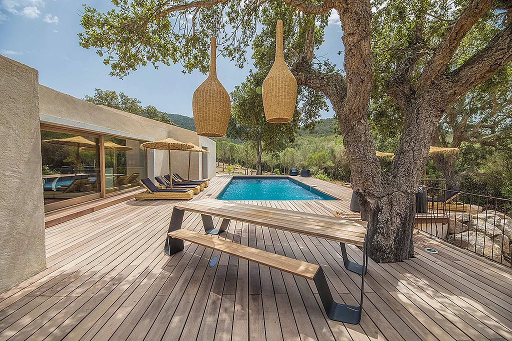 Moderne Betonvilla mit rechteckigem Pool und großzügiger Holzterrasse. Im Vordergrund steht ein hölzerner Hopper-Picknicktisch unter einem großen Baum, über dem zwei Hängelampen hängen.