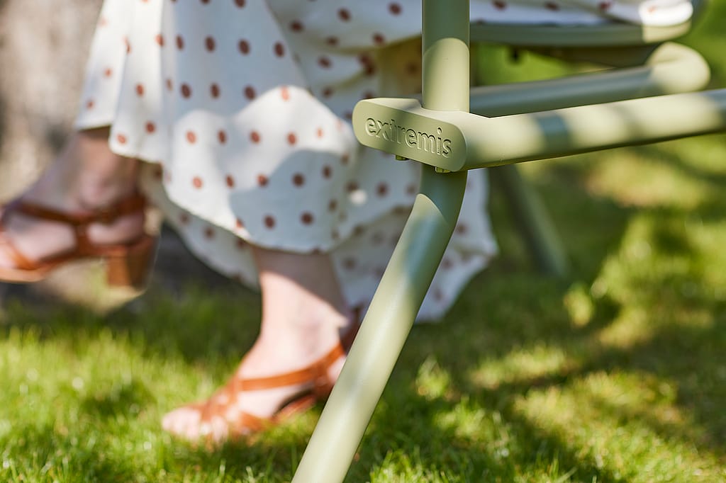 Close-up van het olijfgroene Bistroo-frame en -been met het Extremis-logo, met de jurk en sandalen van een zittende persoon zacht wazig achter.