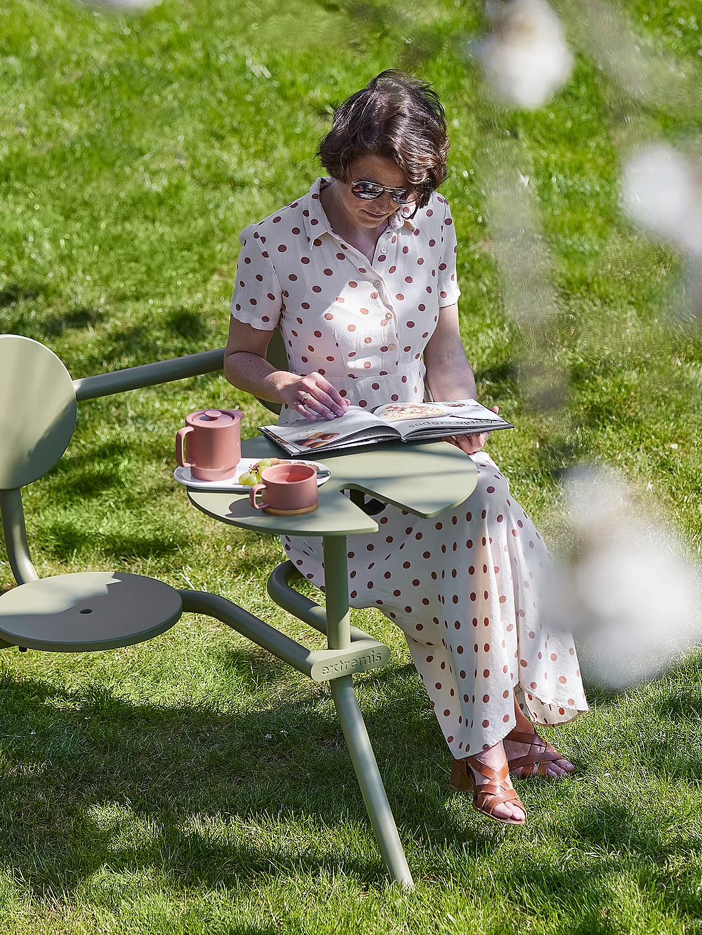 Person sitzt auf einem olivgrünen Bistroo-Stuhl unter einem blühenden Baum und liest eine Zeitschrift mit rosa Tassen und einer kleinen Teekanne auf dem beigefügten Tisch in einem sonnigen Garten.