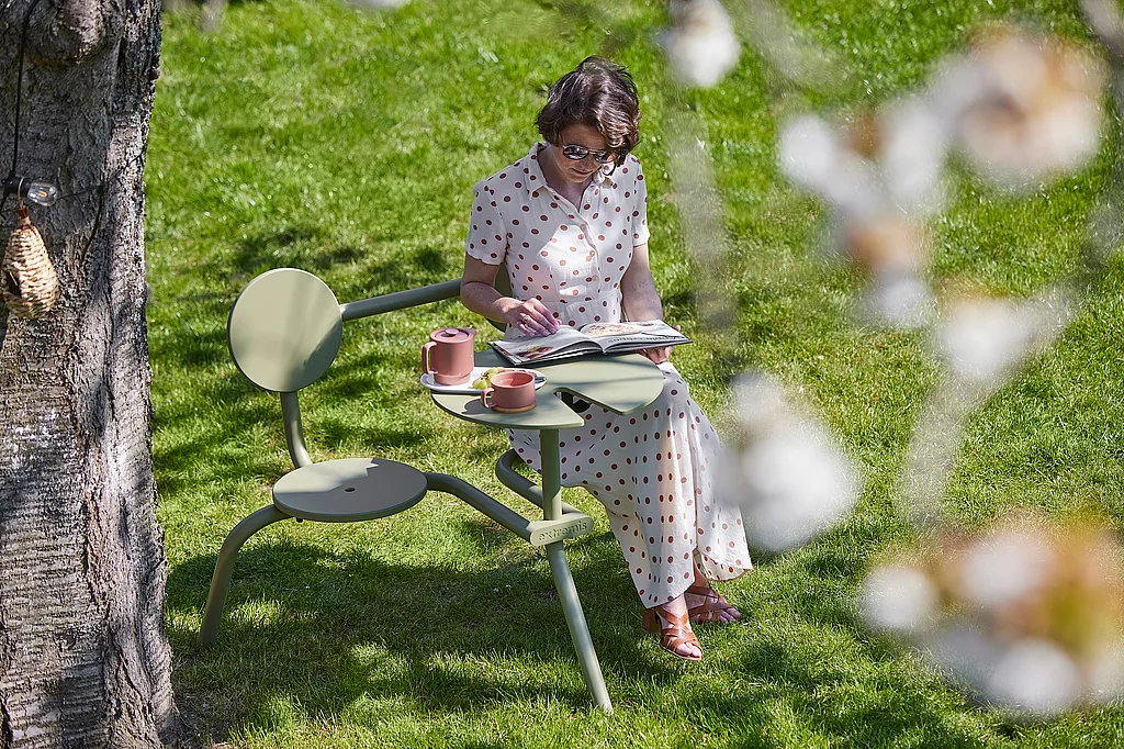 Person sitting on an olive green Bistroo chair under a blossoming tree, reading a magazine with pink cups and a small teapot on the attached tabletop in a sunny garden.