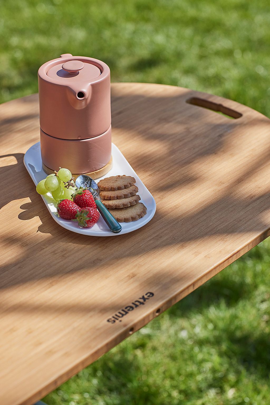 Roze theepot, druiven, aardbeien, lepel en koekjes op een wit dienblad dat buiten op een houten tafelblad wordt geplaatst in gevlekt zonlicht.