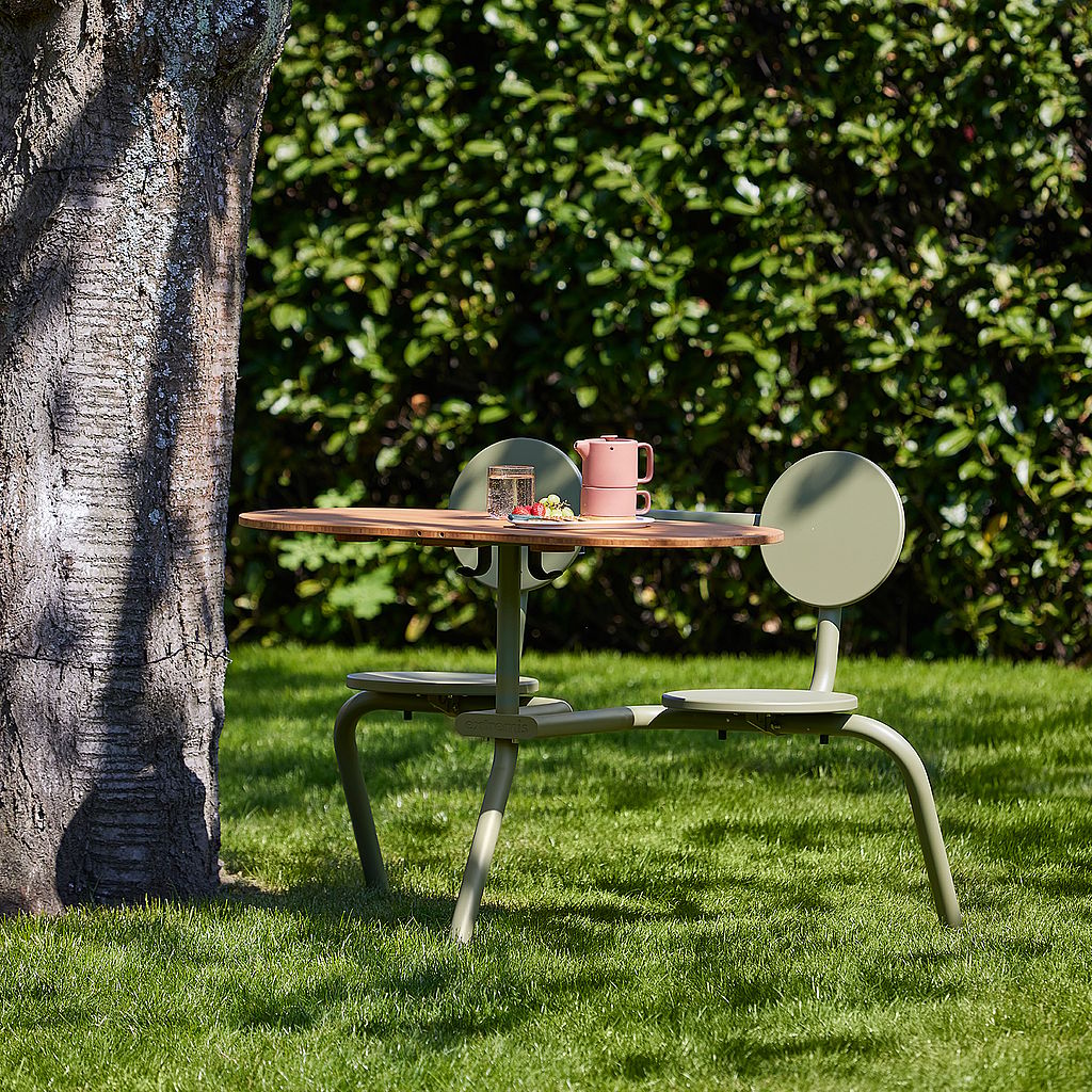 Olijfgroene Bistrootafel en stoelen met een houten tafelbladverlenging op een grasveld naast een grote boom, met een roze theepot, kopjes en snacks bovenop.