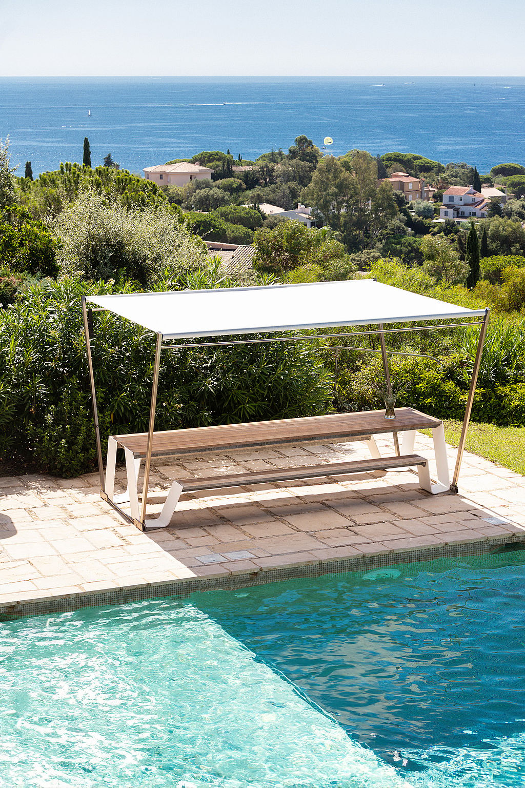 Longue trémie sous une ombre blanche au bord d’une piscine, surplombant une végétation dense, des villas et la mer Méditerranée bleue.