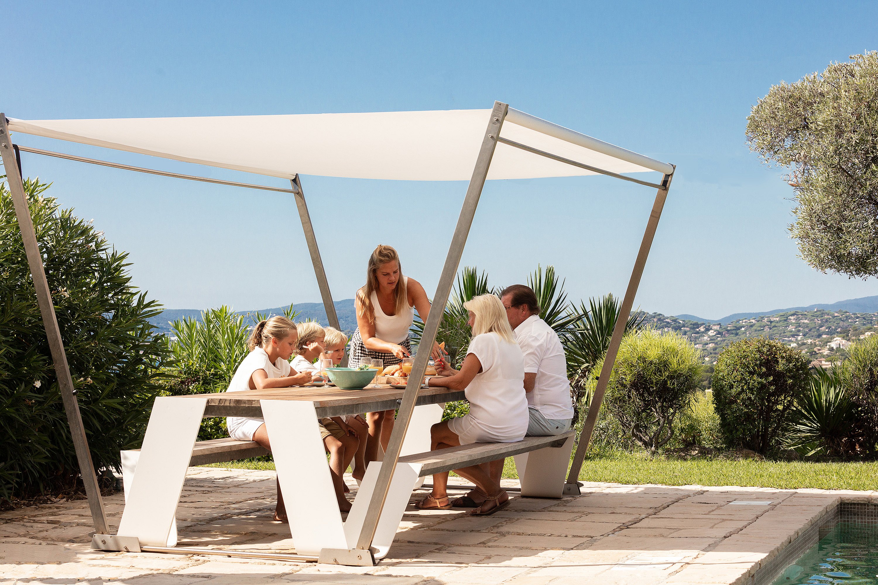 Une famille de cinq personnes s’est réunie autour d’un sauterelle extérieur en bois sous une ombre blanche sur une terrasse en pierre ensoleillée à côté de la piscine, avec des collines côtières au-delà.