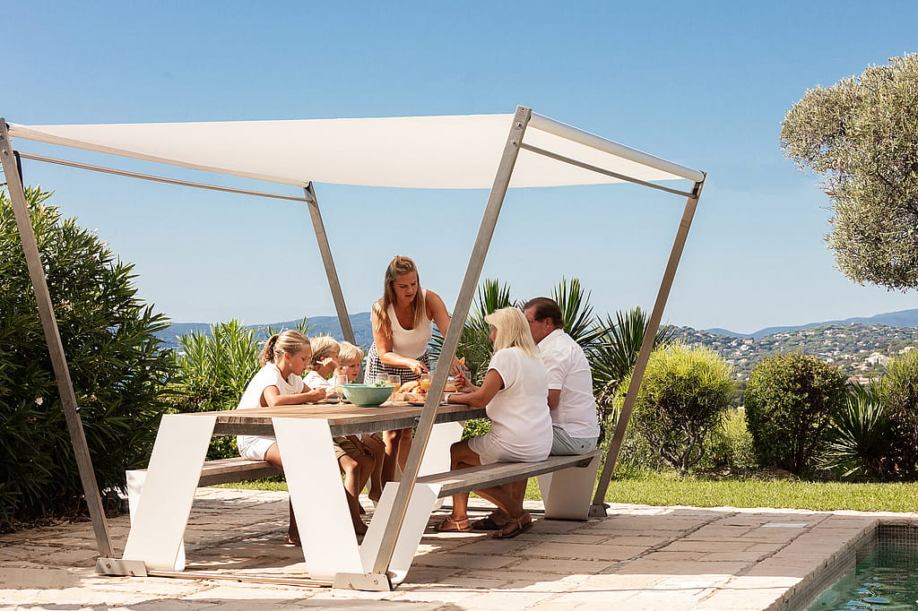 Une famille de cinq personnes s’est réunie autour d’un sauterelle extérieur en bois sous une ombre blanche sur une terrasse en pierre ensoleillée à côté de la piscine, avec des collines côtières au-delà.