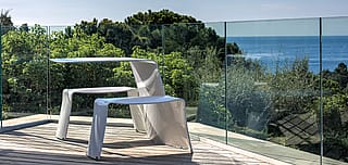 Picnik blanc installé sur un pont en bois à côté d’une balustrade en verre, entouré d’une végétation luxuriante avec la mer au loin.