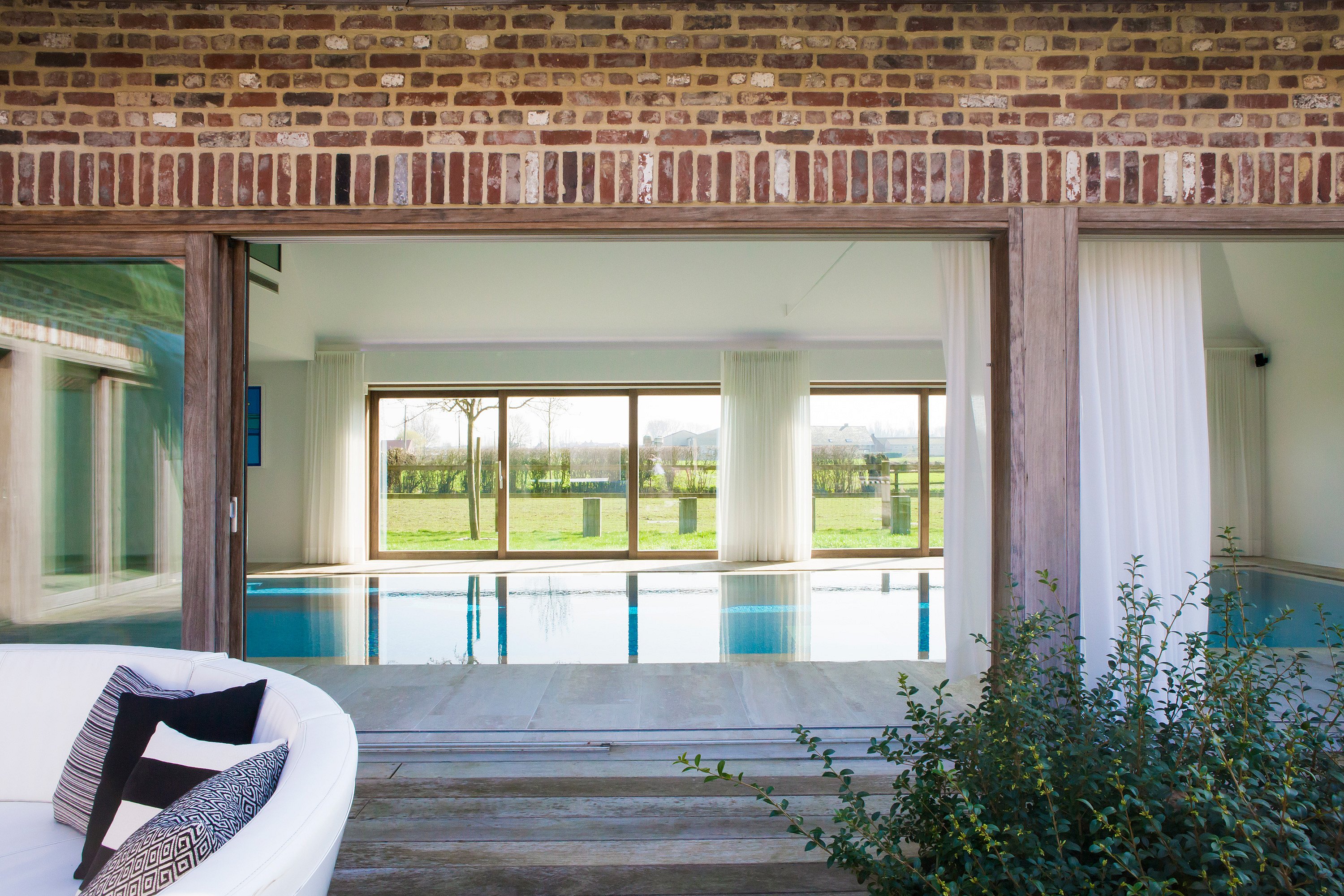   Vue à travers une large façade en briques ouvertes sur une piscine intérieure. Au premier plan, on aperçoit un canapé Kosmos blanc aux courbes harmonieuses et des plantes du jardin.