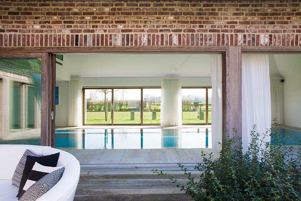Vue à travers une large façade en briques ouvertes sur une piscine intérieure. Au premier plan, on aperçoit un canapé Kosmos blanc aux courbes harmonieuses et des plantes du jardin.