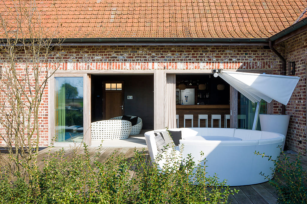 Canapé lounge d'extérieur Kosmos blanc aux courbes harmonieuses, sur une terrasse en bois devant une maison rustique en briques. Un espace de service ouvert et un parasol blanc créent une ambiance décontractée au bord de la piscine.