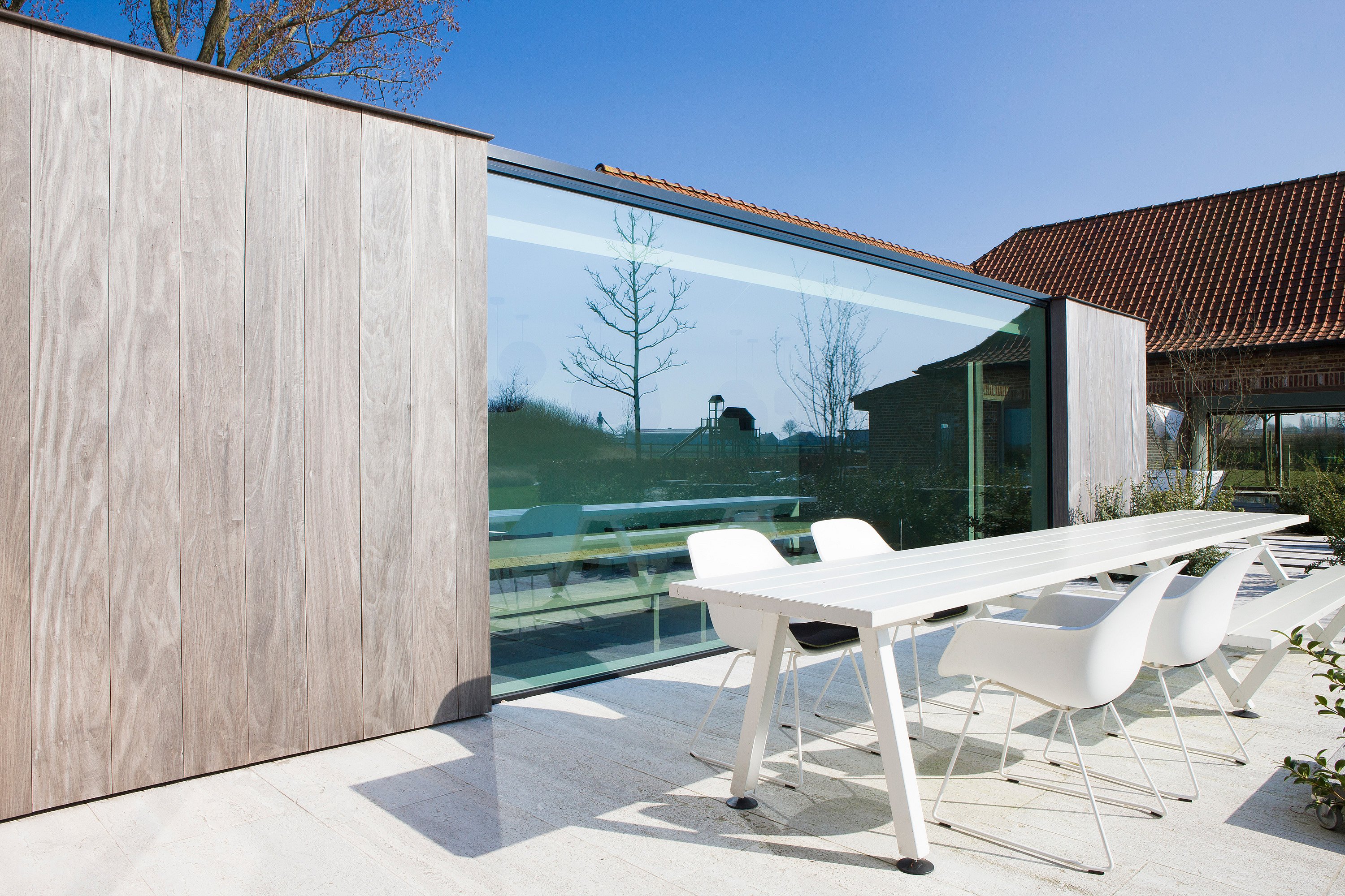 Table de salle à manger extérieure Marina blanche avec chaises blanches assorties, sur une terrasse en pierre claire, jouxtant une extension moderne en verre habillée de bois, sous un ciel d'azur.