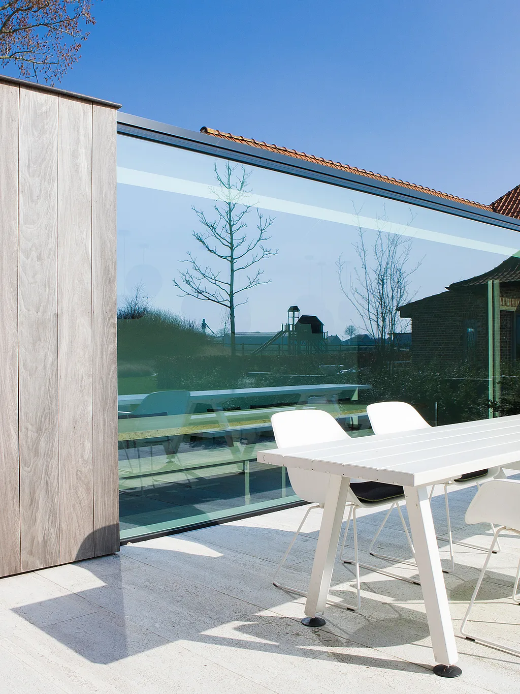 Table de salle à manger extérieure Marina blanche avec chaises blanches assorties, sur une terrasse en pierre claire, jouxtant une extension moderne en verre habillée de bois, sous un ciel d'azur.