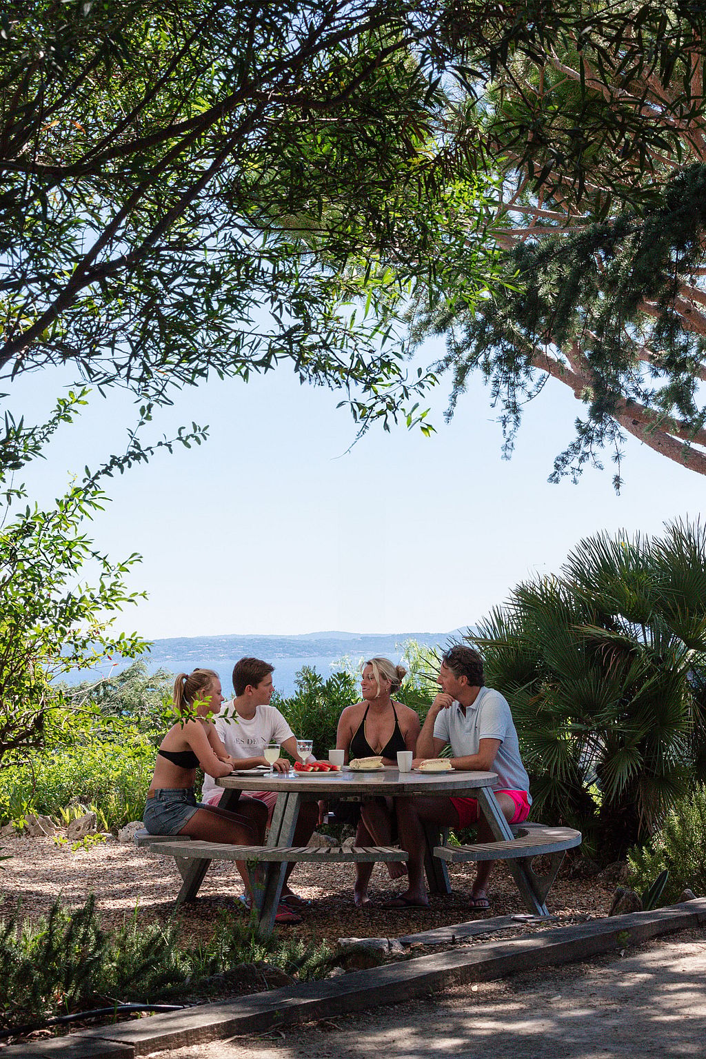 Quatre personnes se sont rassemblées à un gargantuesque sous de grands arbres, encadrées par une végétation dense avec la mer visible au loin.