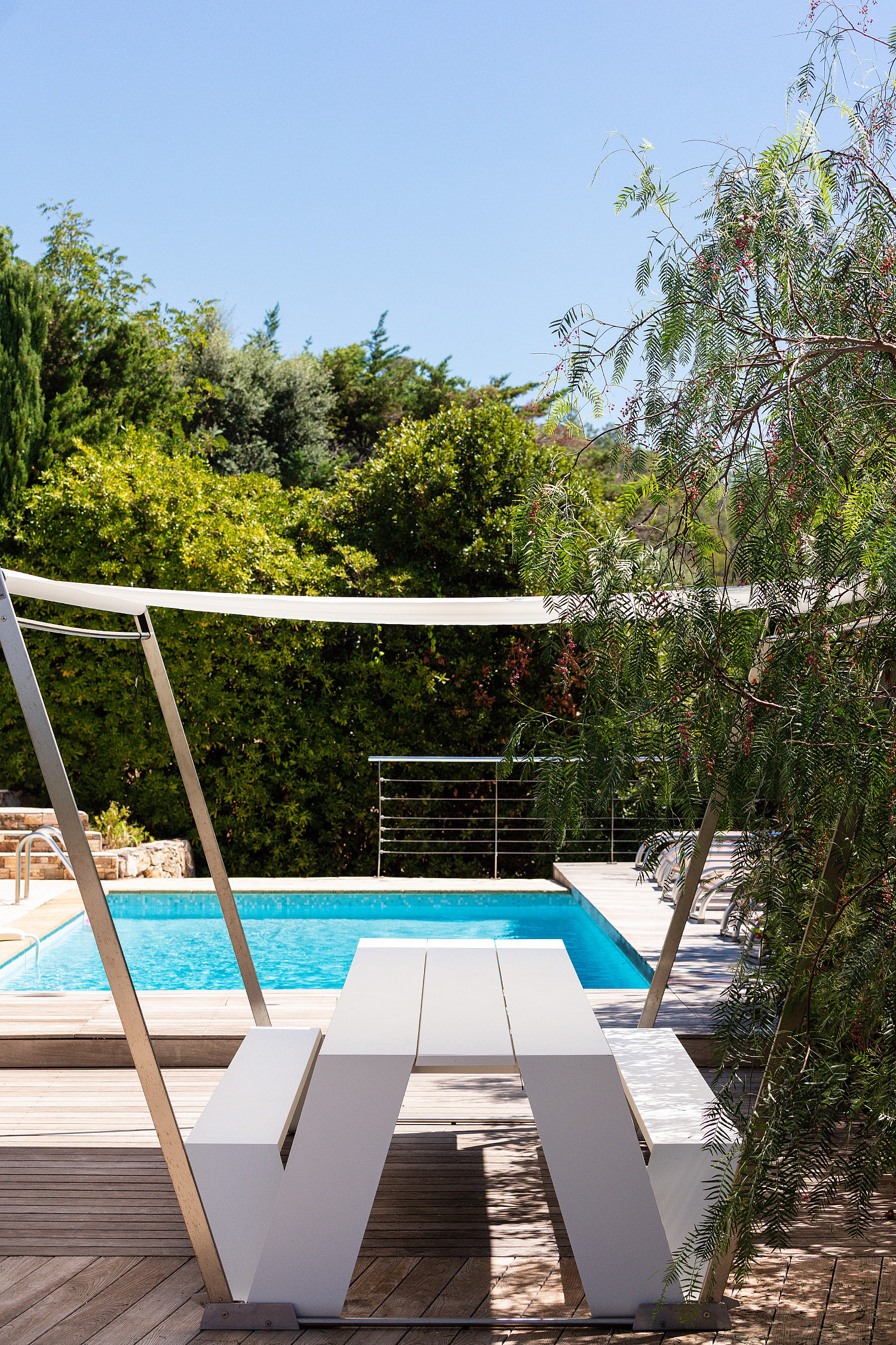 Trémie blanche vide sous une structure d’ombre blanche sur une terrasse en bois à côté de la piscine, bordée d’arbustes feuillus et d’arbres.