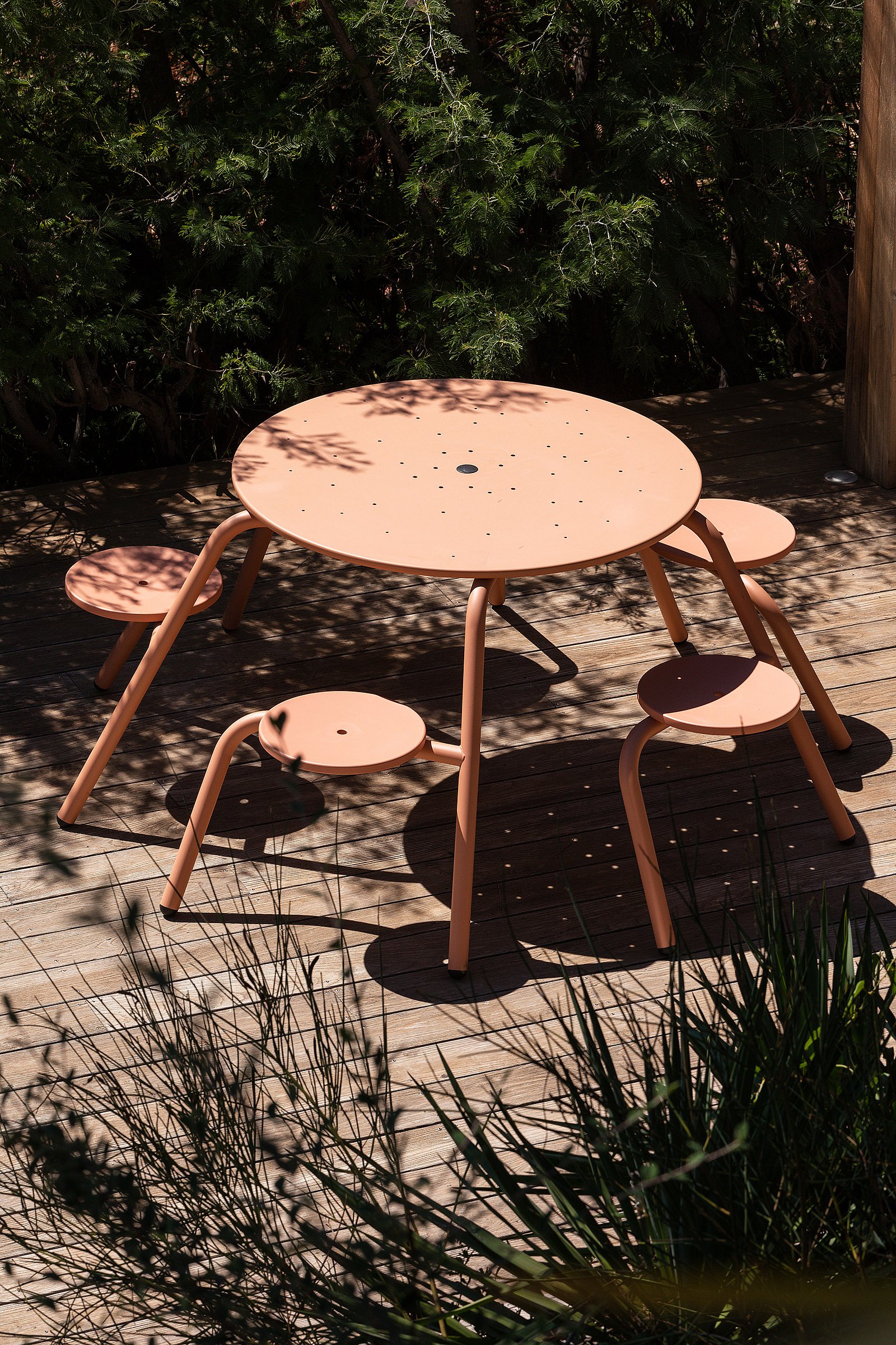 Table de virus rose saumon avec tabourets attachés debout seul sur une terrasse en bois, entourée de plantes et d’ombre tachetée.