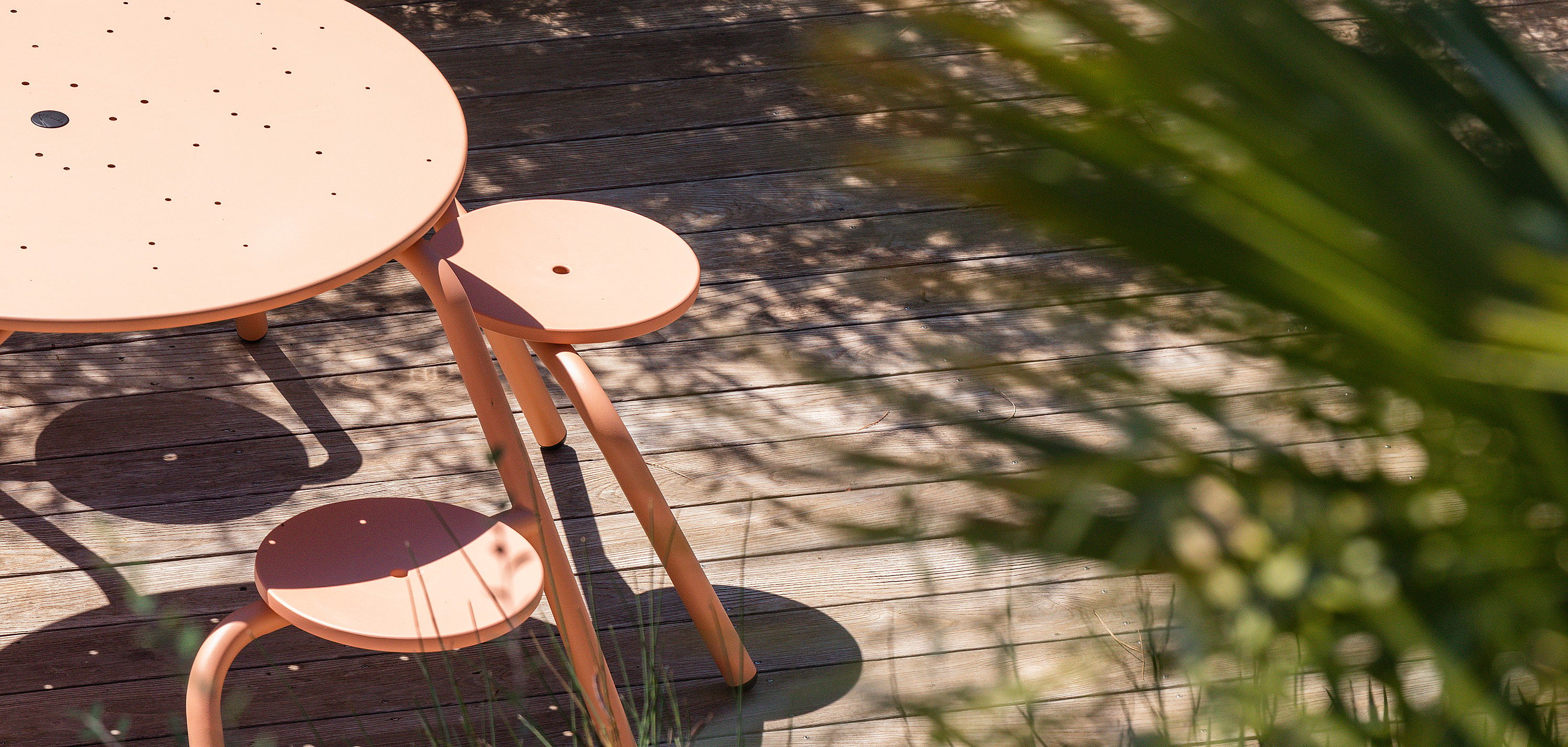 Gros plan partiel d’une table de virus rose saumon avec des tabourets attachés sur une terrasse en bois ensoleillée, avec des ombres de feuilles douces à la surface.