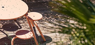 Gros plan partiel d’une table de virus rose saumon avec des tabourets attachés sur une terrasse en bois ensoleillée, avec des ombres de feuilles douces à la surface.