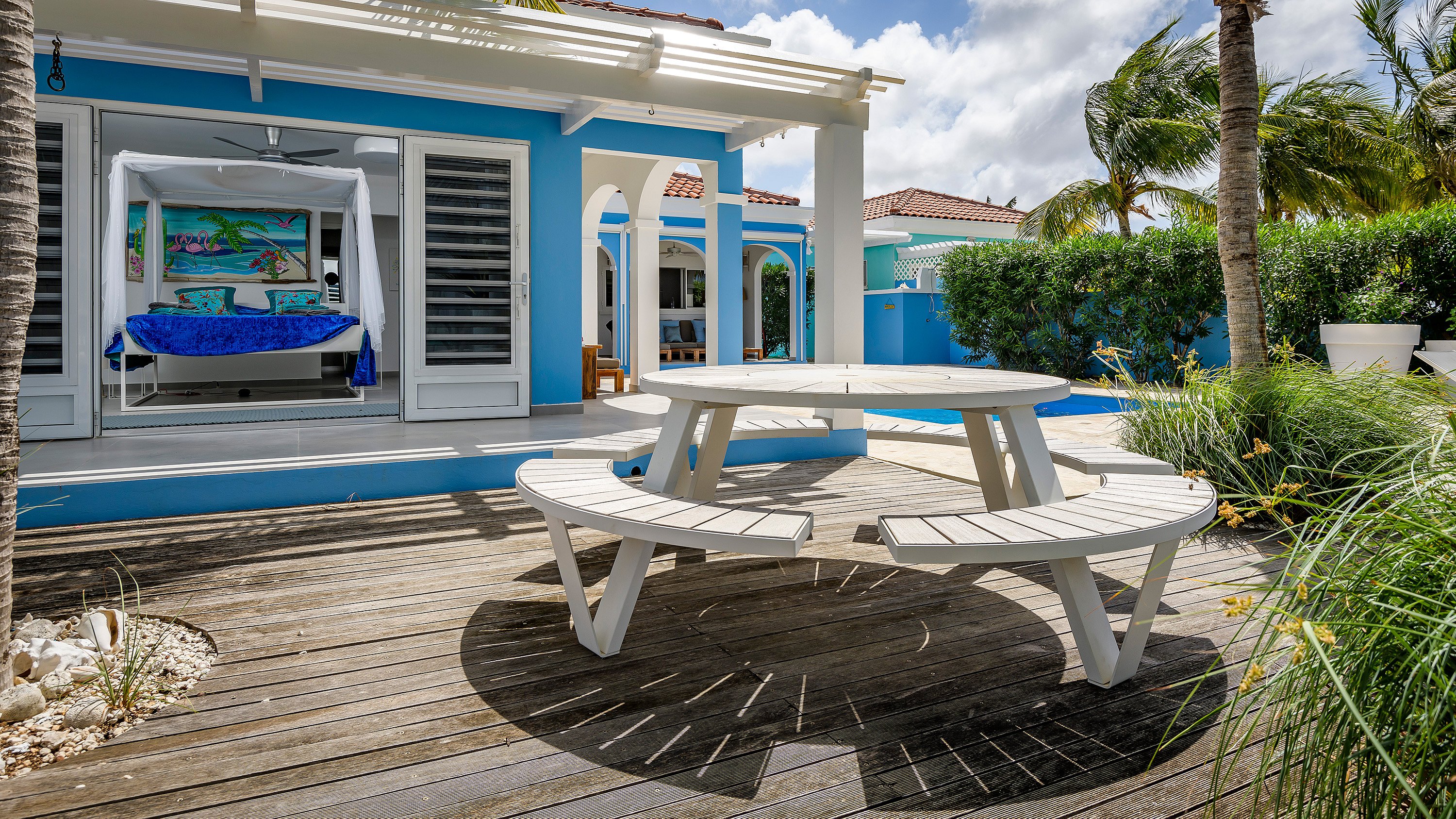 White round Pantagruel picnic table on a wooden deck outside a bright blue tropical villa, with white arches, palm trees, and an open bedroom visible through sliding doors.