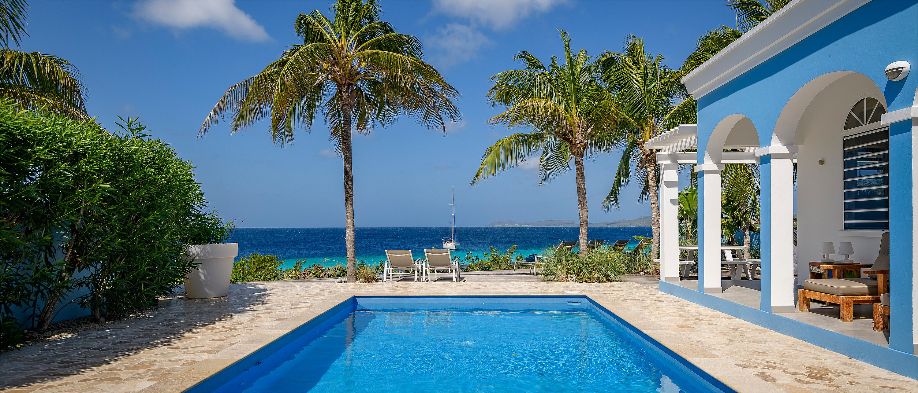 lue beach villa with white arches beside a rectangular swimming pool, overlooking palm trees, lounge chairs, and a turquoise sea with a sailboat in the distance.
