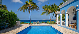lue beach villa with white arches beside a rectangular swimming pool, overlooking palm trees, lounge chairs, and a turquoise sea with a sailboat in the distance.