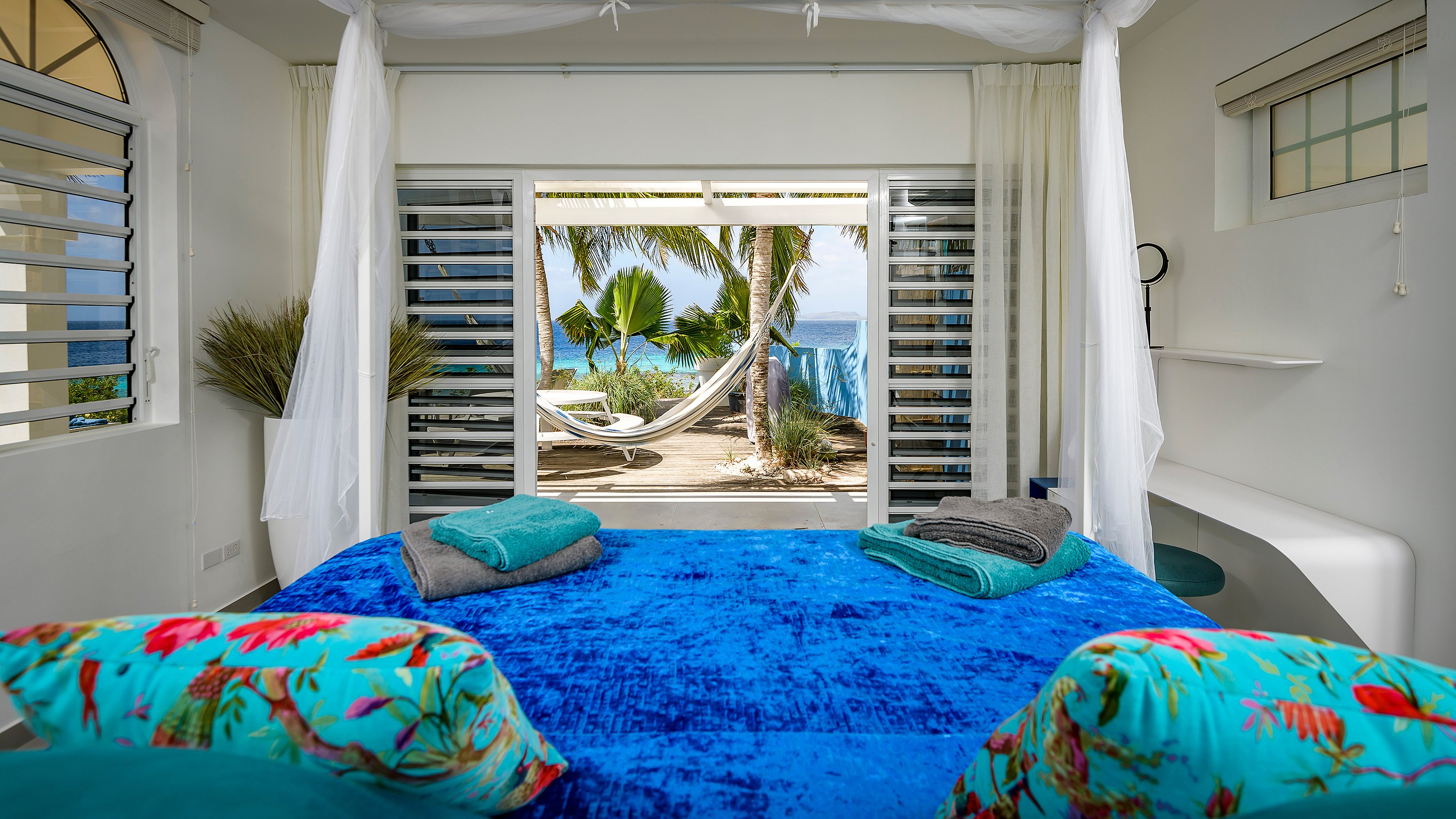 Bright white bedroom with a blue bedspread and floral pillows, opening through sliding doors onto a terrace with a hammock, palm trees, and a view of the sea.