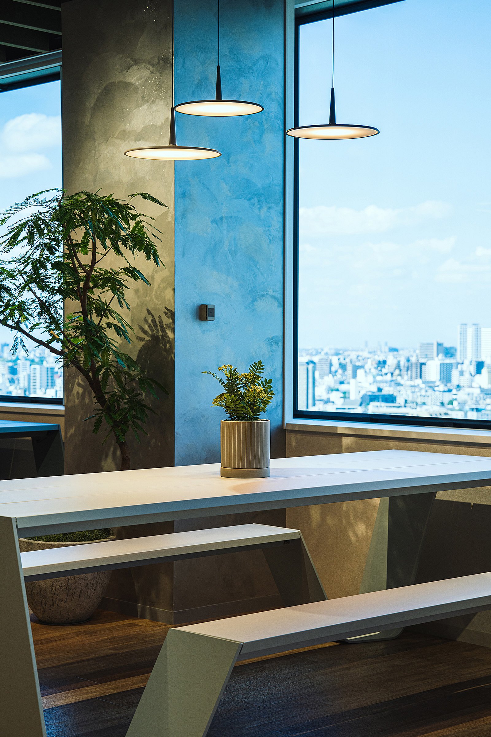 Close-up of a white Hopper table and bench beside a floor-to-ceiling window, with three pendant lights, potted plants, and a bright city view beyond.