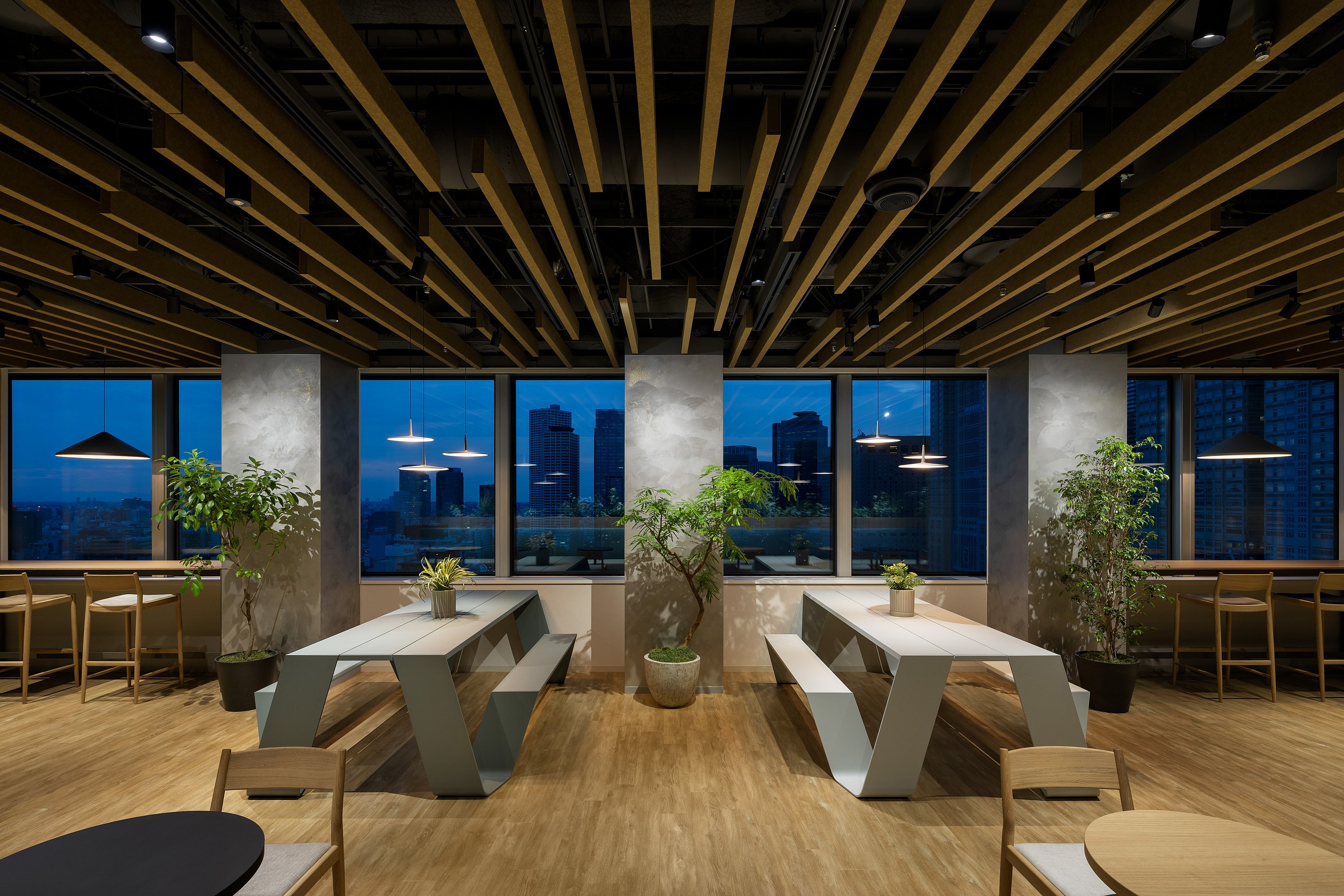 Modern office lounge with two white Hopper picnic tables, potted trees, and pendant lights, set in front of large windows overlooking the Tokyo skyline at dusk.