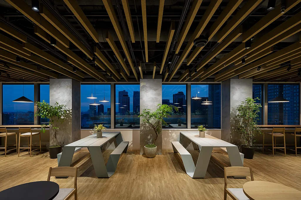 Modern office lounge with two white Hopper picnic tables, potted trees, and pendant lights, set in front of large windows overlooking the Tokyo skyline at dusk.