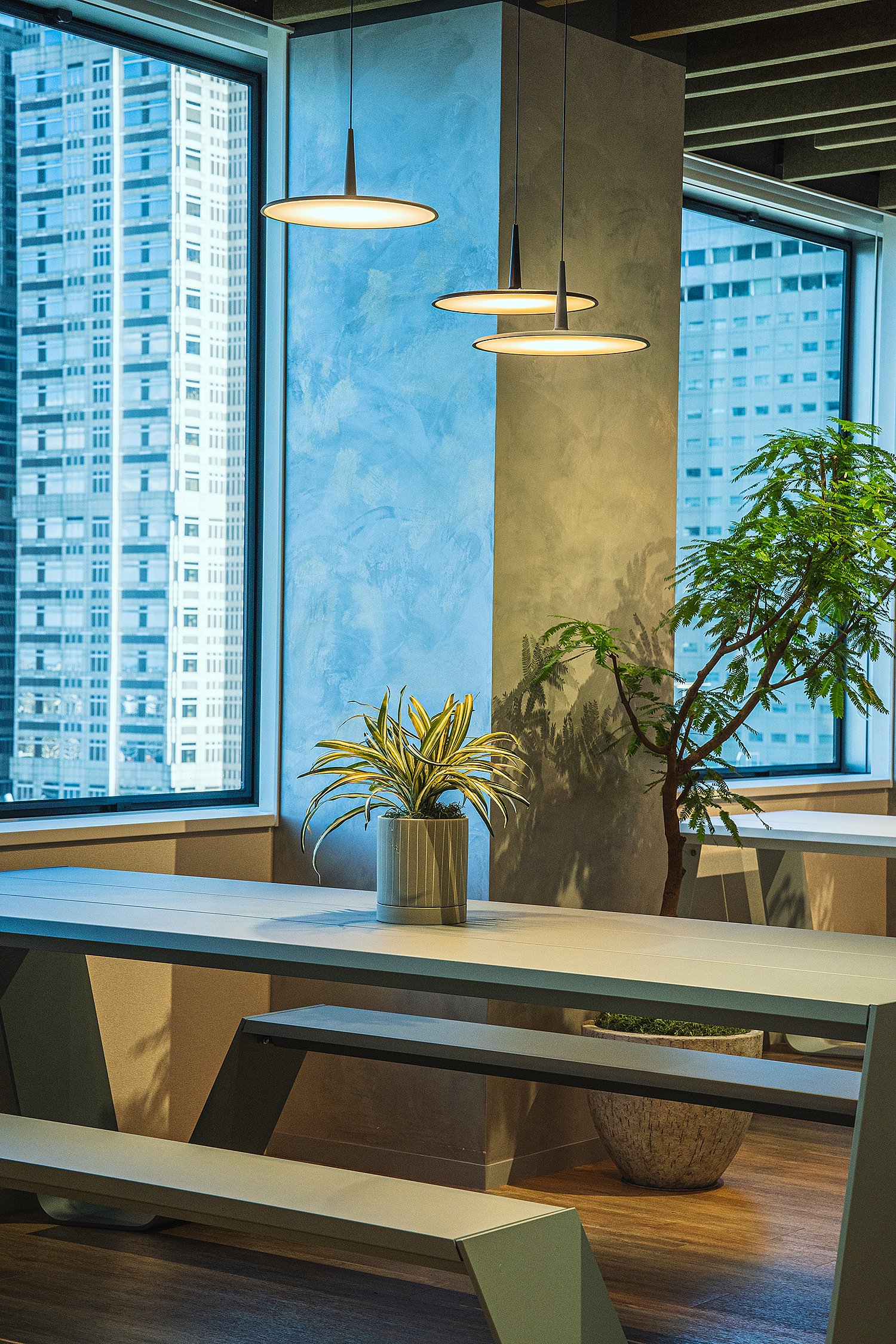 Close-up of a white Hopper table and bench beside a floor-to-ceiling window, with three pendant lights, potted plants, and a bright city view beyond.