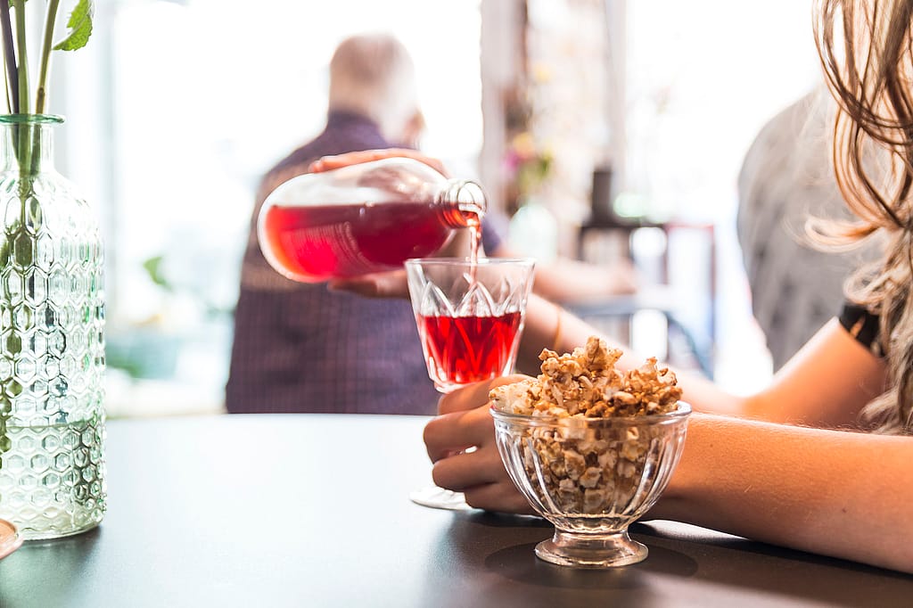 Nahaufnahme von rotem Tee, der in ein Glas auf einer dunklen Tischplatte gegossen wird, mit einer kleinen Glasschüssel Müsli und sanft verschwommenen Bäckereigästen im Hintergrund.