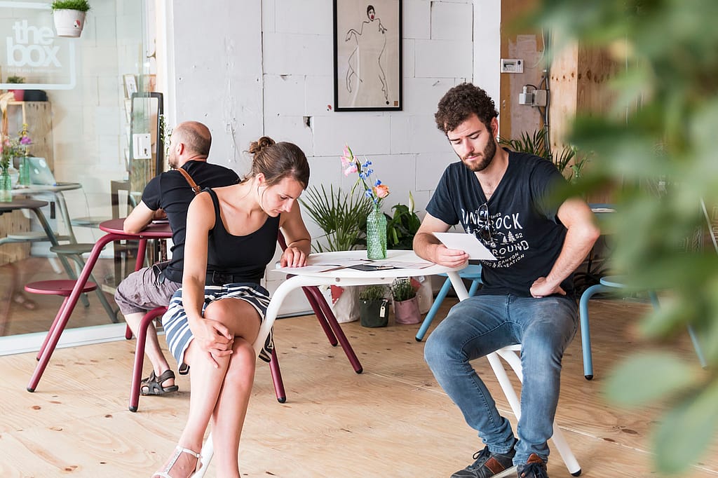 Junger Mann und junge Frau sitzen an getrennten weißen Virus-Picknicktischen auf einem Holzboden und lesen Menüs in der hellen Pop-up-Bäckerei.