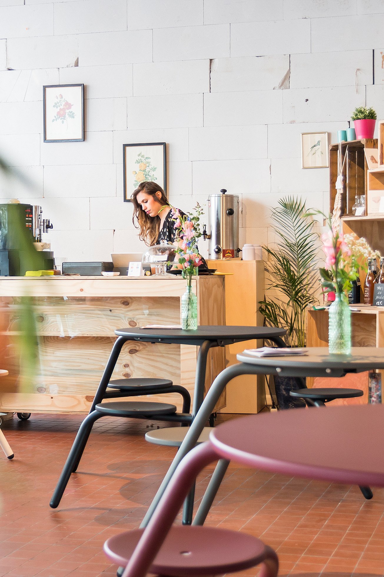 Intérieur chaleureux avec des tables de pique-nique pastel Virus au premier plan, tandis qu’une femme travaille derrière un comptoir en bois entouré de fleurs, d’étagères et de réfrigérateurs à présentoirs.