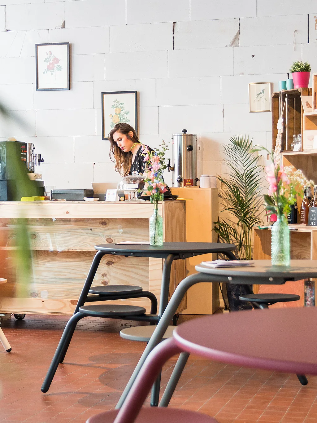 Intérieur chaleureux avec des tables de pique-nique pastel Virus au premier plan, tandis qu’une femme travaille derrière un comptoir en bois entouré de fleurs, d’étagères et de réfrigérateurs à présentoirs.