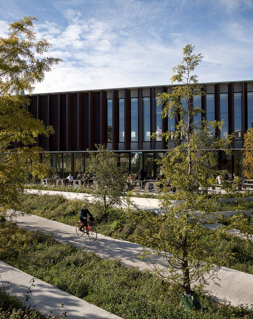 Cyclist riding along a winding path through lush planting beds in front of a long dark building façade, with outdoor seating tucked among the greenery.