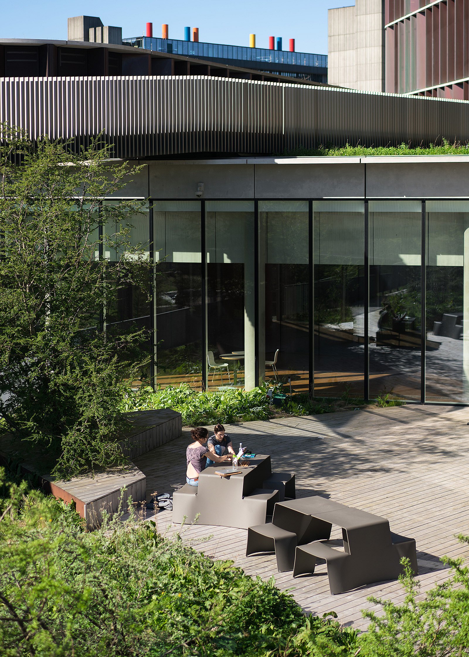 Open campus plaza with several long picnik tables in front of the tall Mærsk Tower, surrounded by young trees and planted swales.