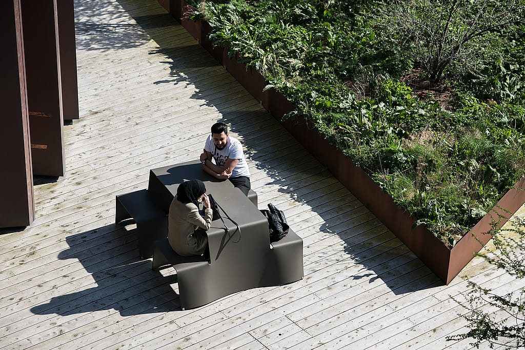 People sitting at picnik tables along a broad paved path through the landscaped park, with the modern campus buildings behind them.