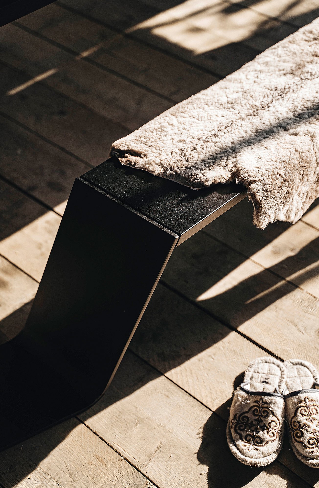 Black picnic table beside a bench draped with a cream sheepskin, lit by warm sunlight inside the cabin, with a cornfield outside the glass wall.