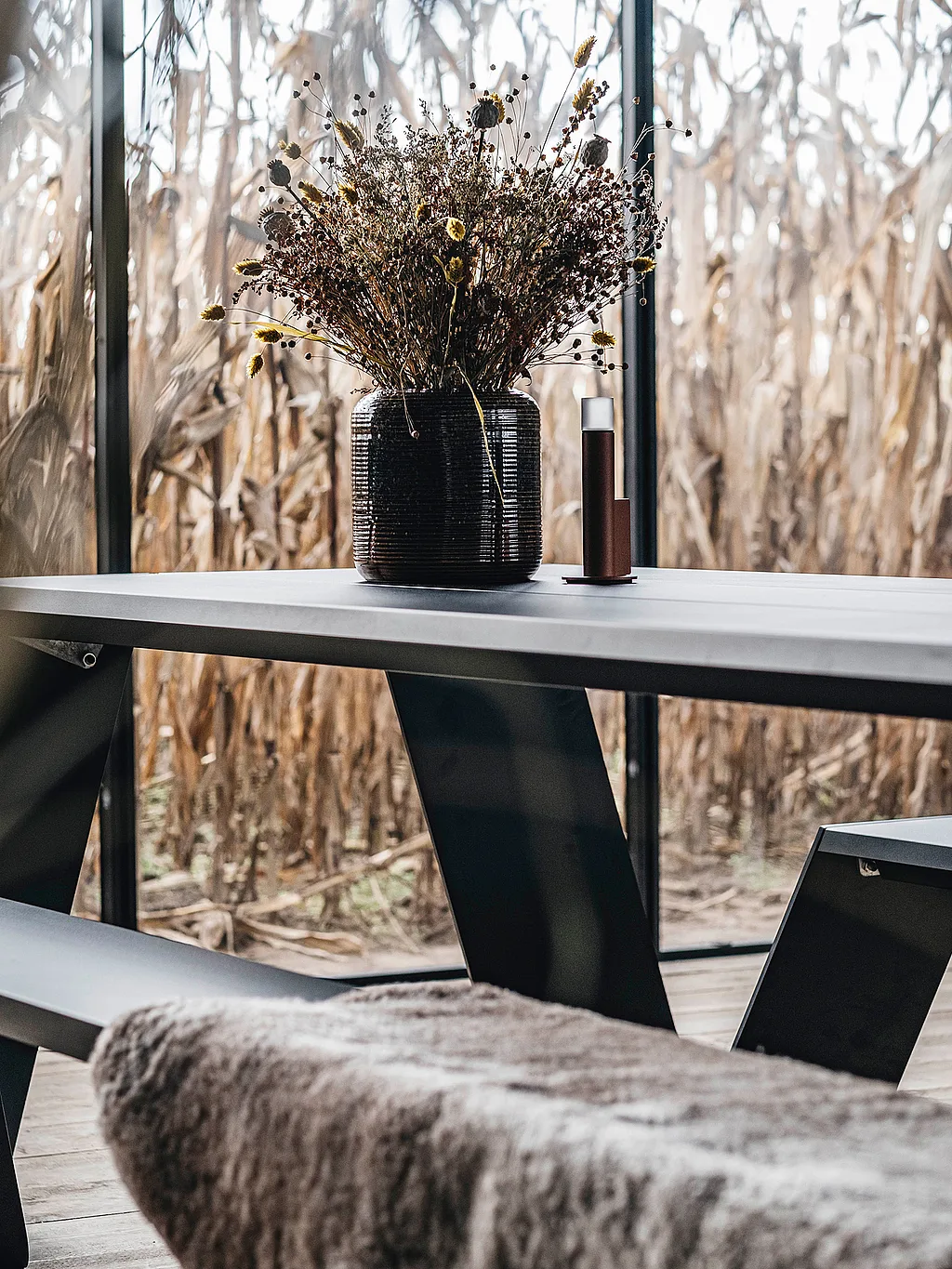 Zwarte Hopper picknicktafel op een houten vloer in een hut met glazen wanden, met gedroogde bloemen op het tafelblad en hoge korenvelden zichtbaar door de kamerhoge ramen.