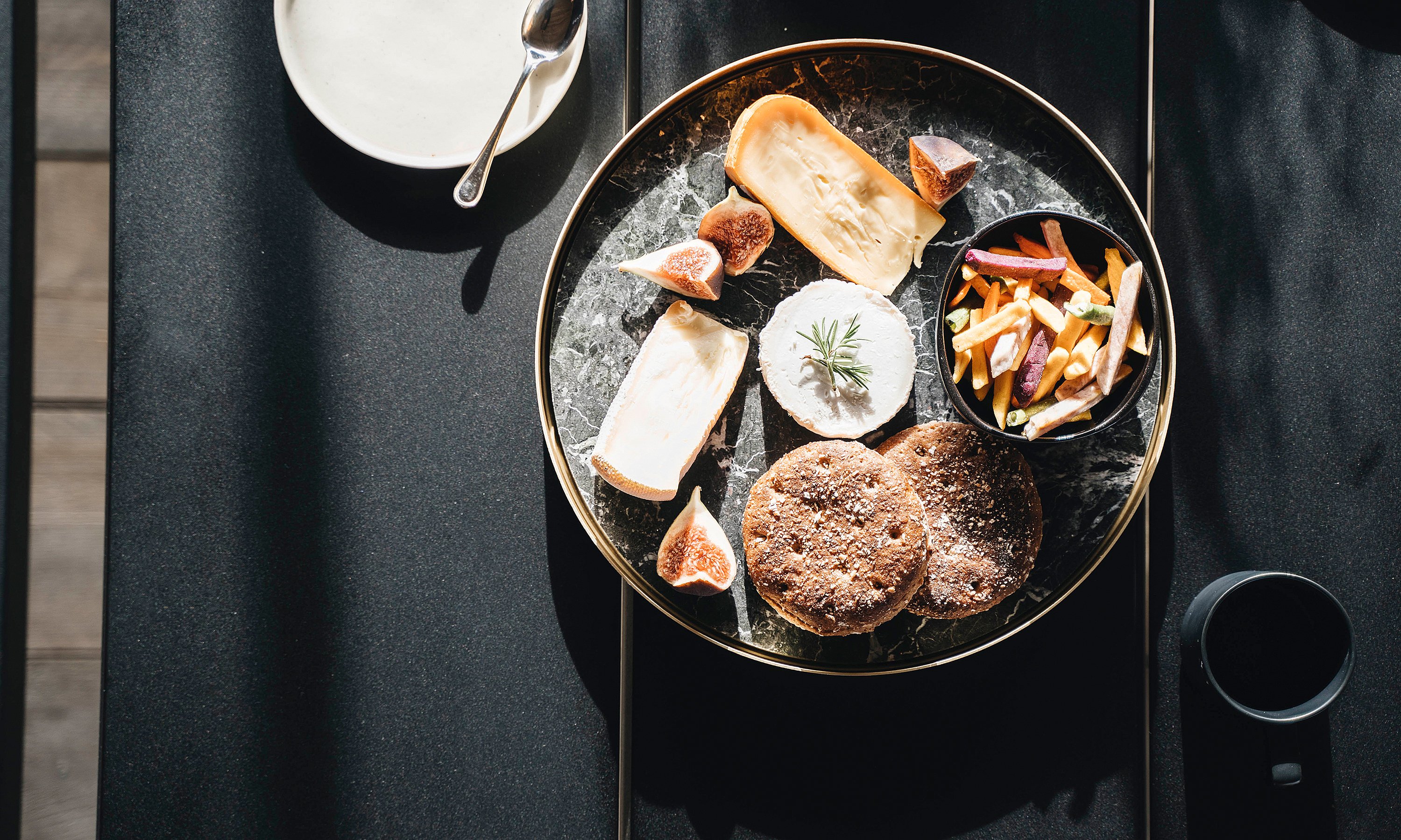 Overhead view of a round serving tray with bread, cheese, figs, and vegetable fries on a black tabletop, with a dark mug beside it.