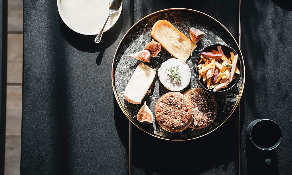 Overhead view of a round serving tray with bread, cheese, figs, and vegetable fries on a black tabletop, with a dark mug beside it.