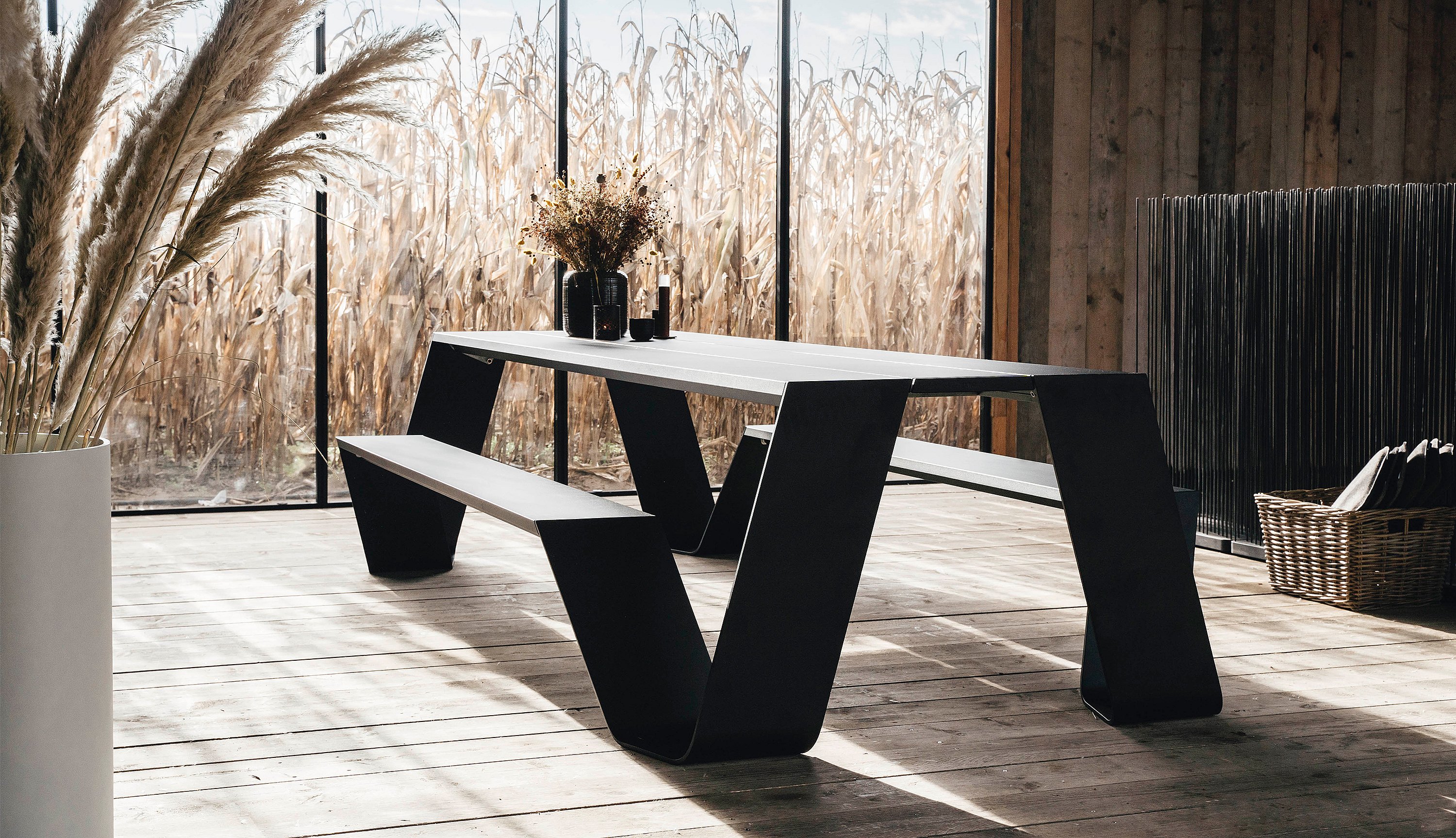 Wide view of the black Hopper picnic table inside the wooden cabin, framed by dried grasses, a basket of slippers, and golden cornfields outside the windows.
