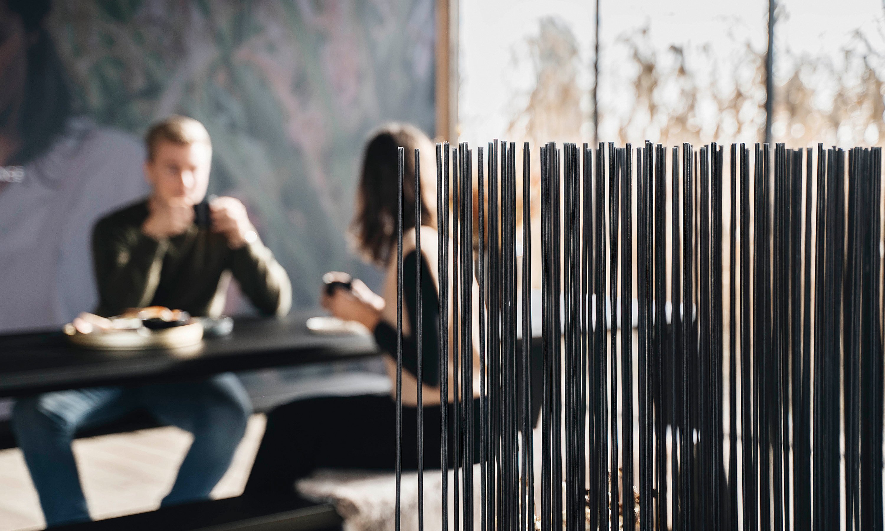 Black Sticks room divider in sharp focus, with two people seated and dining at the table in the softly blurred background.