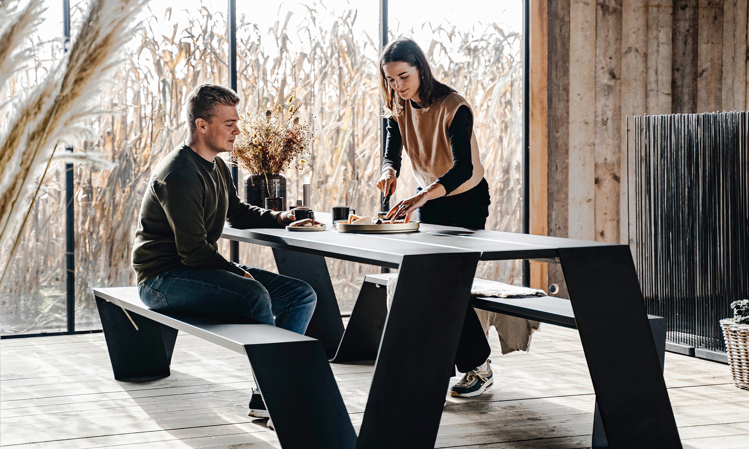 Man seated at a black Hopper picnic table while a woman serves food, inside a cozy timber cabin surrounded by cornfields seen through large windows.