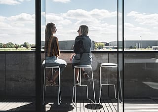 Drei weiße Tiki-Barhocker aufgereiht auf einem Balkon hinter einer hohen Glasfassade, mit Blick auf eine offene Landschaft unter einem Himmel mit vereinzelten Wolken.