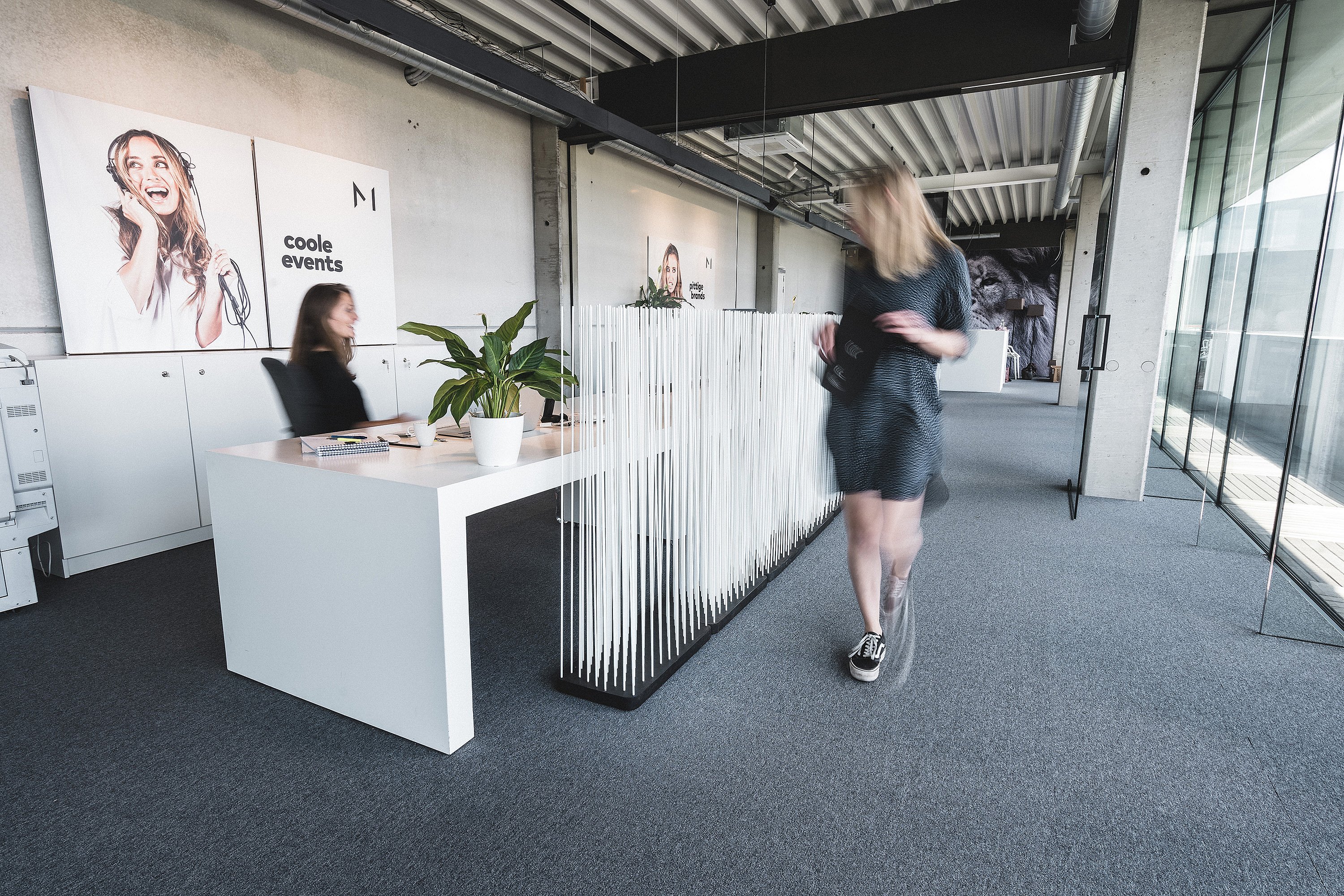 White Sticks-Raumteiler neben einer Reihe weißer Schreibtische in einem hellen Bürokorridor im Industriestil, mit einer Person, die an gläsernen Besprechungsräumen vorbeigeht.