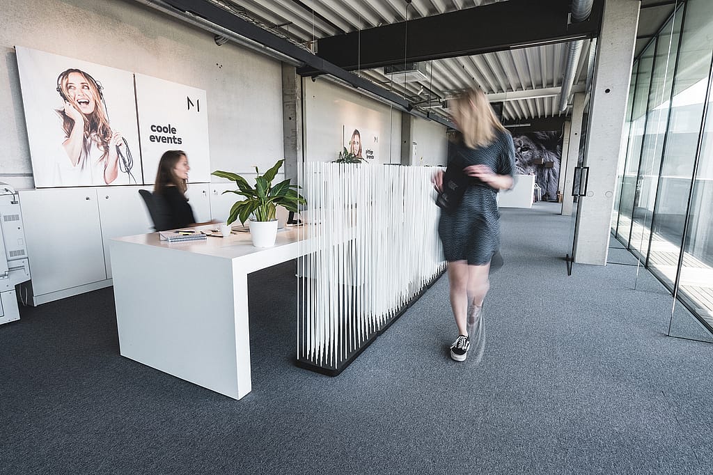 White Sticks-Raumteiler neben einer Reihe weißer Schreibtische in einem hellen Bürokorridor im Industriestil, mit einer Person, die an gläsernen Besprechungsräumen vorbeigeht.
