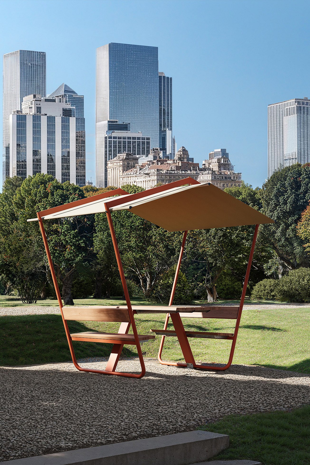 Wide elevated view of a city park with three roofed Manille picnic tables on gravel and decking, surrounded by lawns, trees, paths, and a skyline of tall towers in the background.
