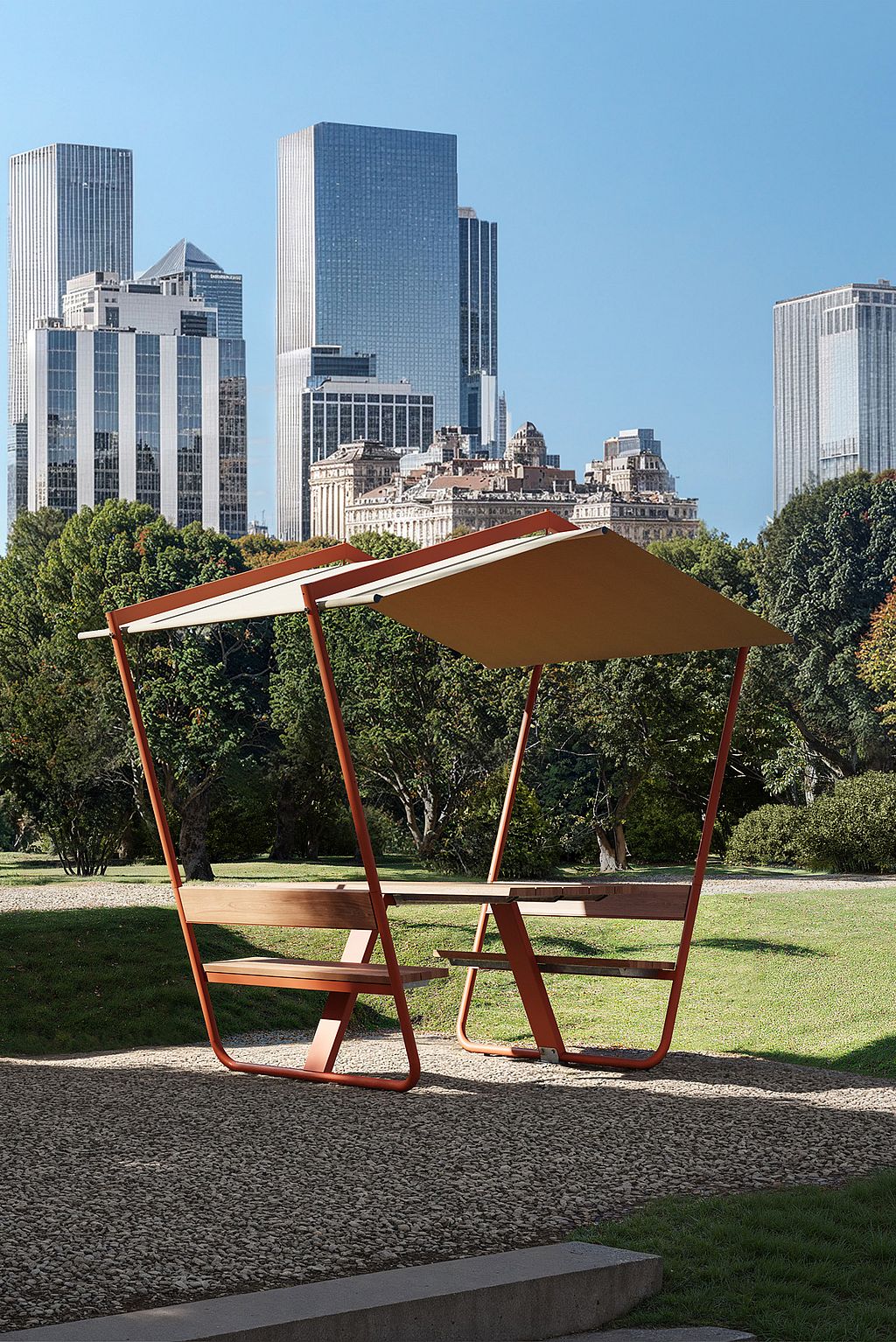 Wide elevated view of a city park with three roofed Manille picnic tables on gravel and decking, surrounded by lawns, trees, paths, and a skyline of tall towers in the background.