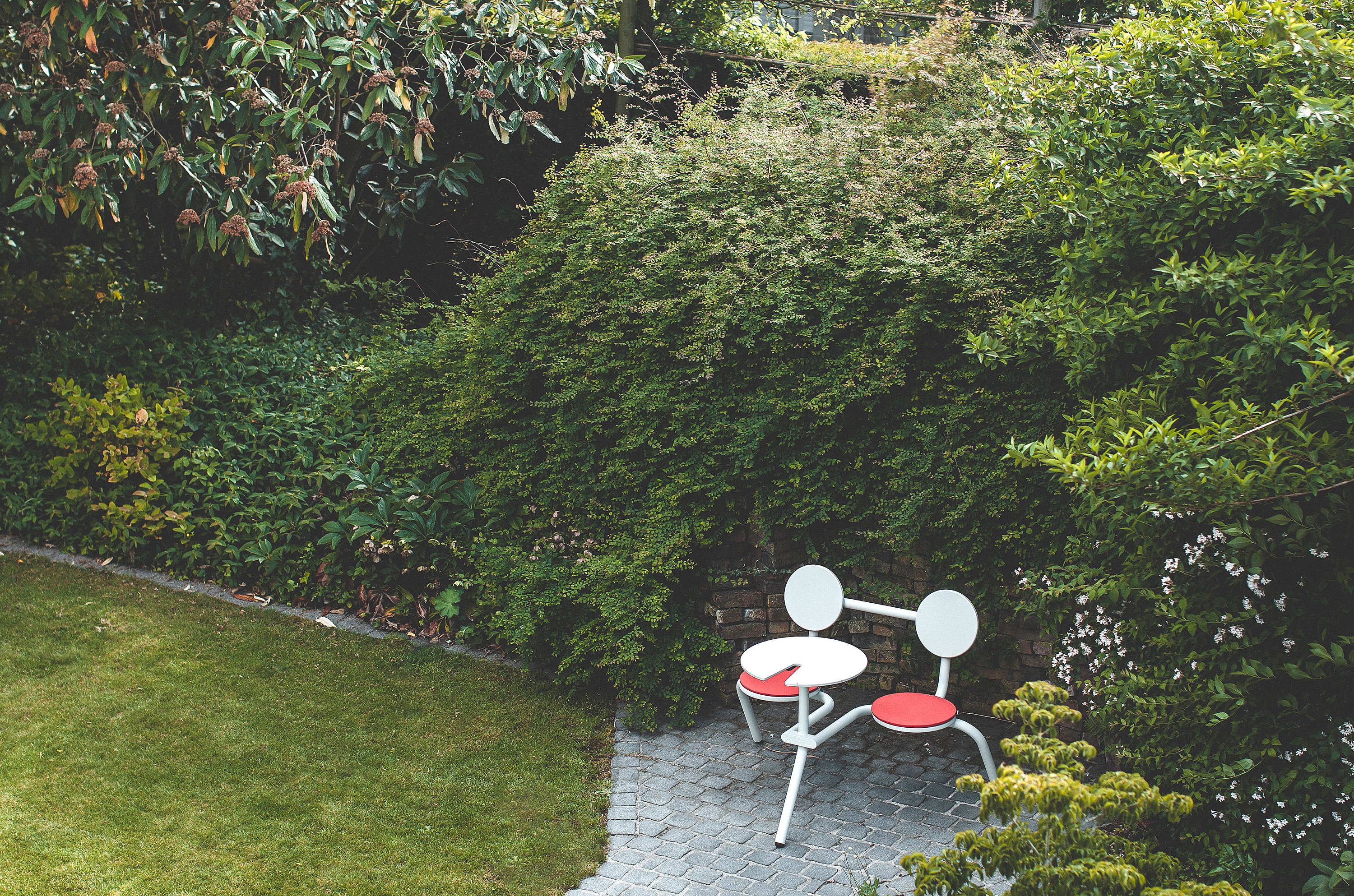 Light gray two-seat outdoor bistro table with round backs and coral-red seat cushions, tucked into a garden corner beside a curved cobblestone path and dense flowering shrubs.