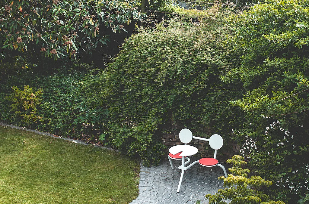 Light gray two-seat outdoor bistro table with round backs and coral-red seat cushions, tucked into a garden corner beside a curved cobblestone path and dense flowering shrubs.