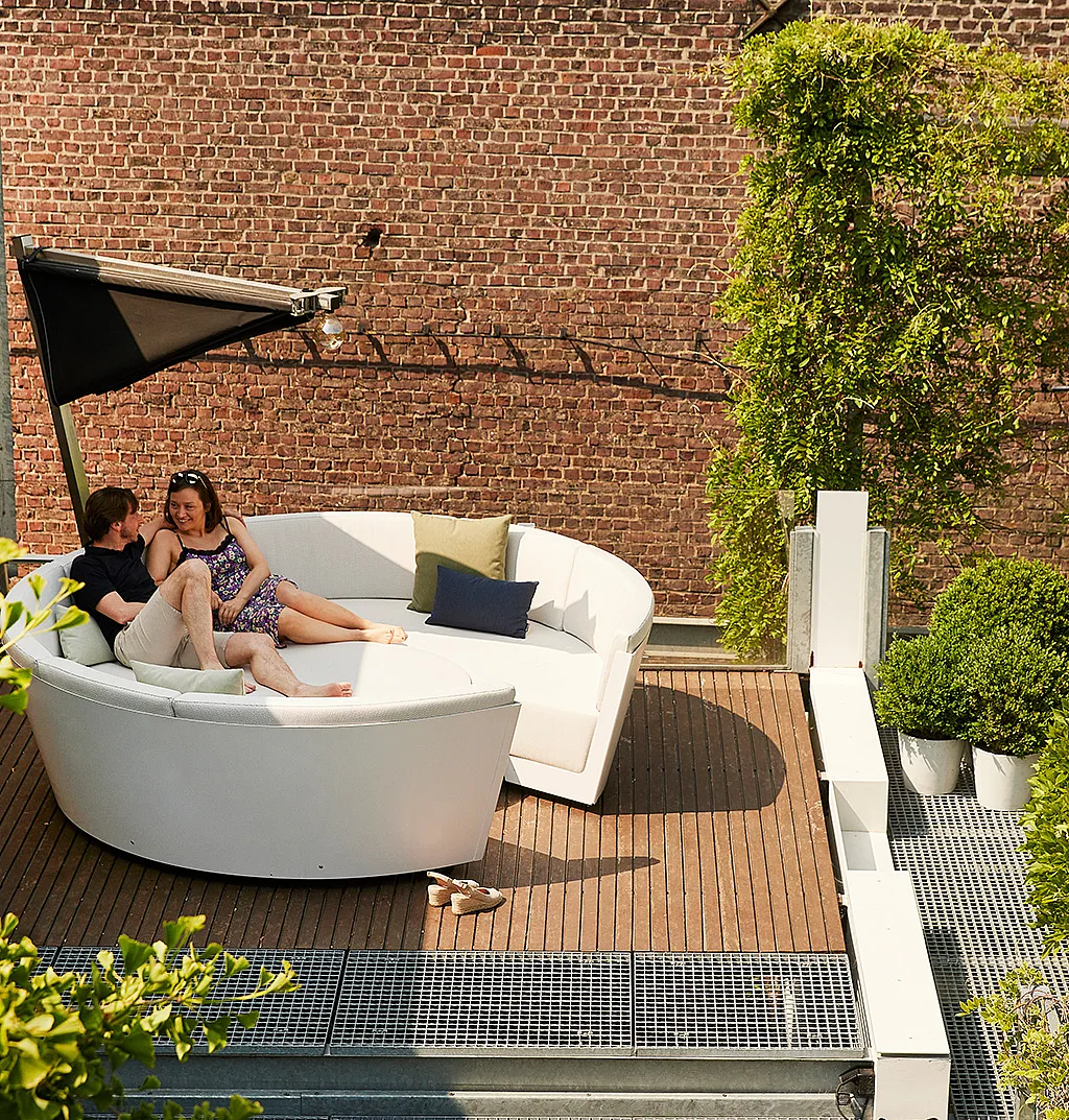 White Kosmos lounge on a wooden rooftop deck, where a couple sit together beneath a black folding shade beside a brick wall, climbing greenery, and a glimpse of a blue pool.
